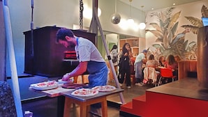 Un chef prepara pizzas en una cocina, con un comedor visible al fondo.