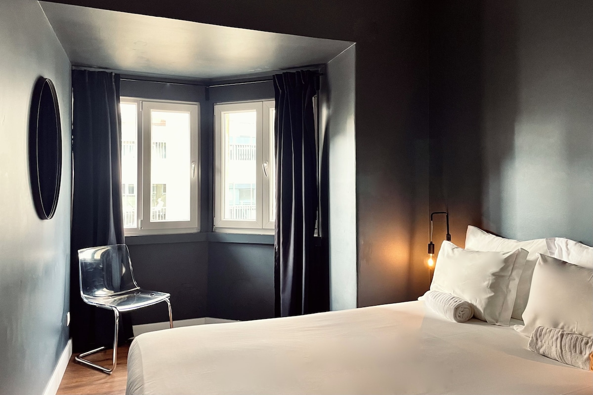 A modern hotel room with a large bed, a chair, two windows with curtains, and a wall-mounted light fixture.