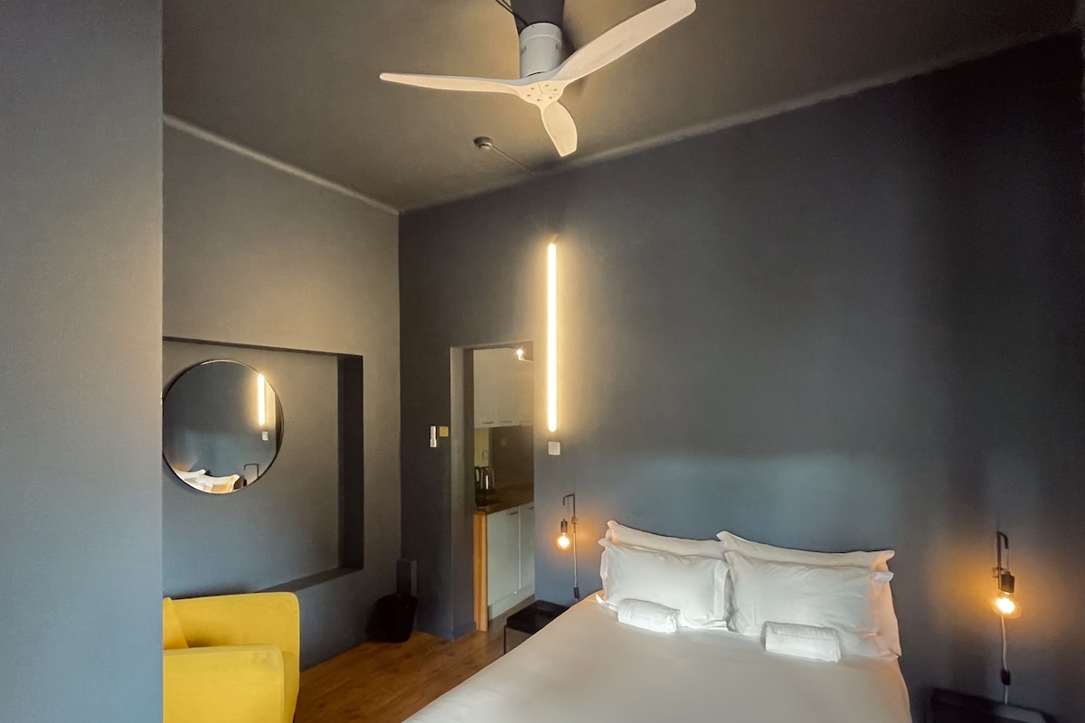 Egy modern szállodai szoba, amelyben egy nagy ágy, egy mennyezeti ventilátor, egy kerek tükör és egy sárga szék található.