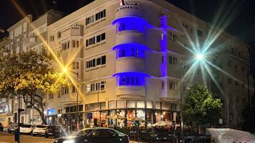 Un hotel de varios pisos con fachada curva, iluminado por luces azules y amarillas, y una cafetería en la planta baja.