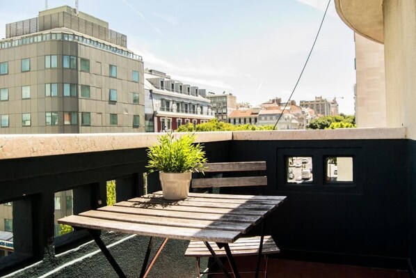 A balcony with a wooden table and a potted plant, overlooking a cityscape with modern buildings. - Avenue Rooms & Suites - Hostel (Lisbon)