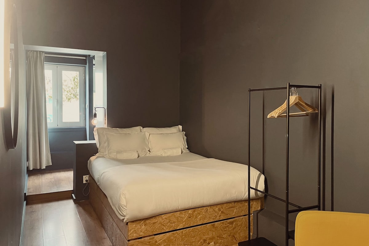 A bedroom with a bed, a wooden floor, a window with curtains, a bedside table, and a clothing rack.