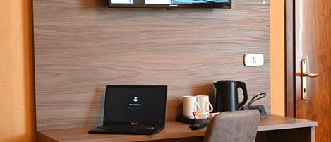 A hotel room with a wooden desk, a laptop, a television mounted on the wall, a chair, a bed with a red cover, and a door.