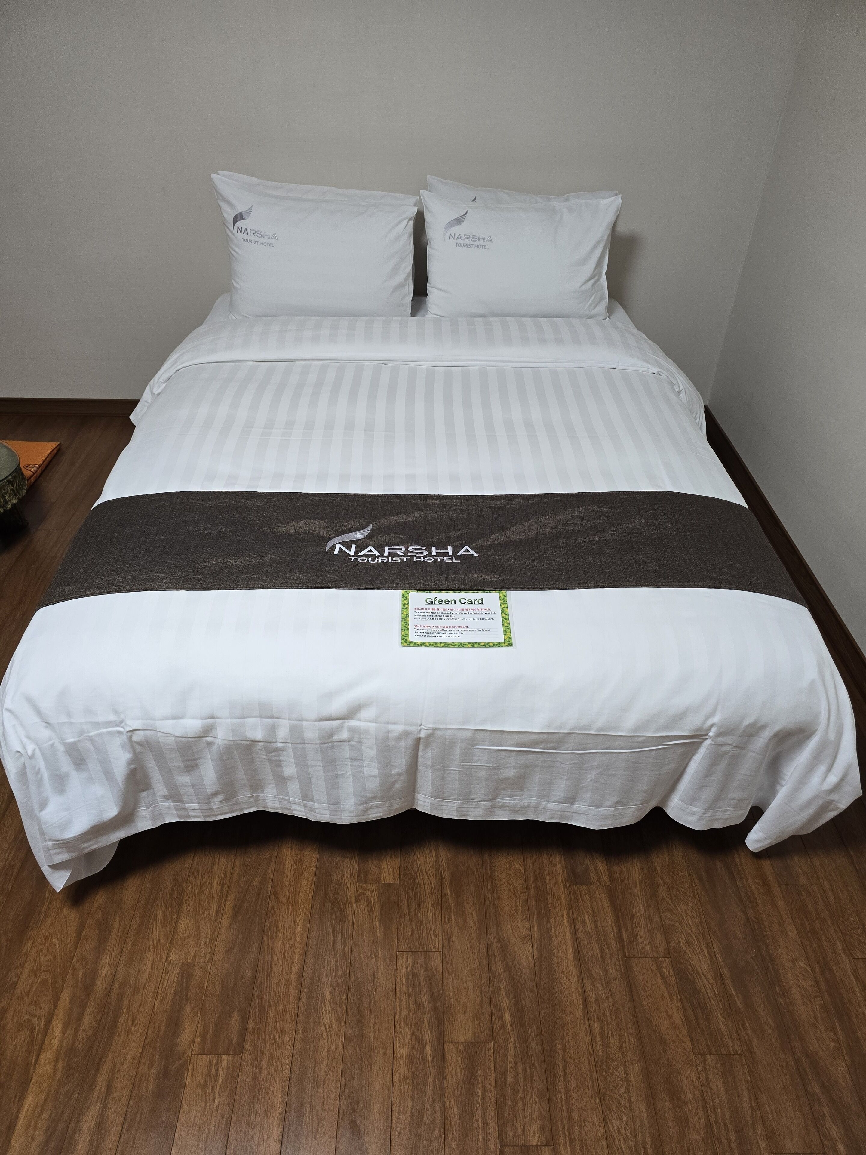 A neatly made bed with white bedding and a dark grey blanket, a green card on the bed, and wooden flooring.