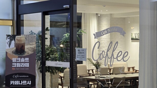 Ein Café mit Glastür, Innenbestuhlung und einem Schild mit der Aufschrift 'Cafe Narsha Coffee'.