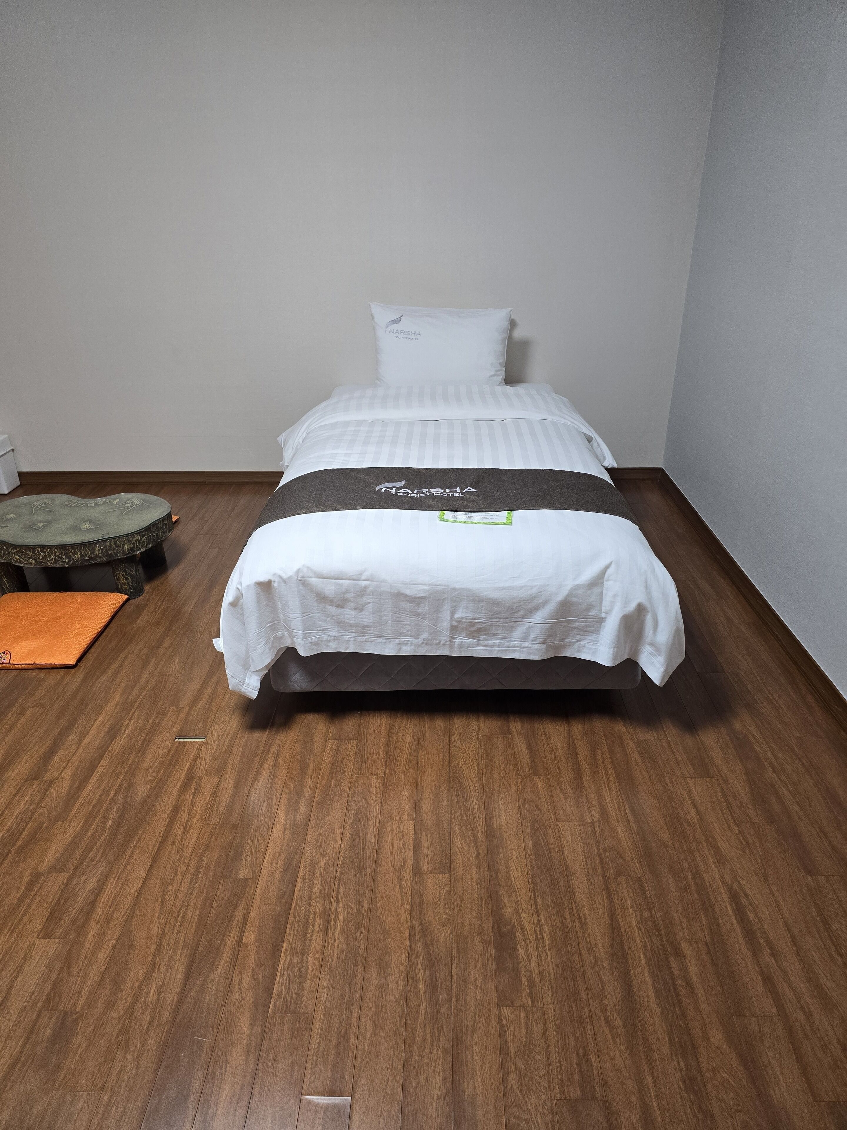 Una cama individual con colcha blanca y gris a rayas, un almohadón con la marca 'NARSHA' y una manta a juego. A la izquierda, hay un pequeño taburete con un cojín verde y otro naranja sobre el suelo. La habitación tiene suelo de madera y paredes blancas.