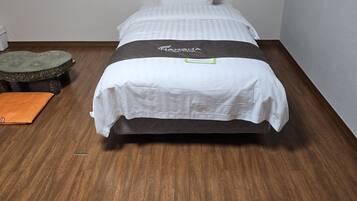 Una cama individual con colcha blanca y gris a rayas, un almohadón con la marca 'NARSHA' y una manta a juego. A la izquierda, hay un pequeño taburete con un cojín verde y otro naranja sobre el suelo. La habitación tiene suelo de madera y paredes blancas.