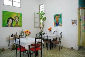 Comedor con una mesa dispuesta para cuatro personas, rodeada de sillas con cojines rojos. Hay dos cuadros en la pared, una ventana con marco de metal y una planta en el centro.