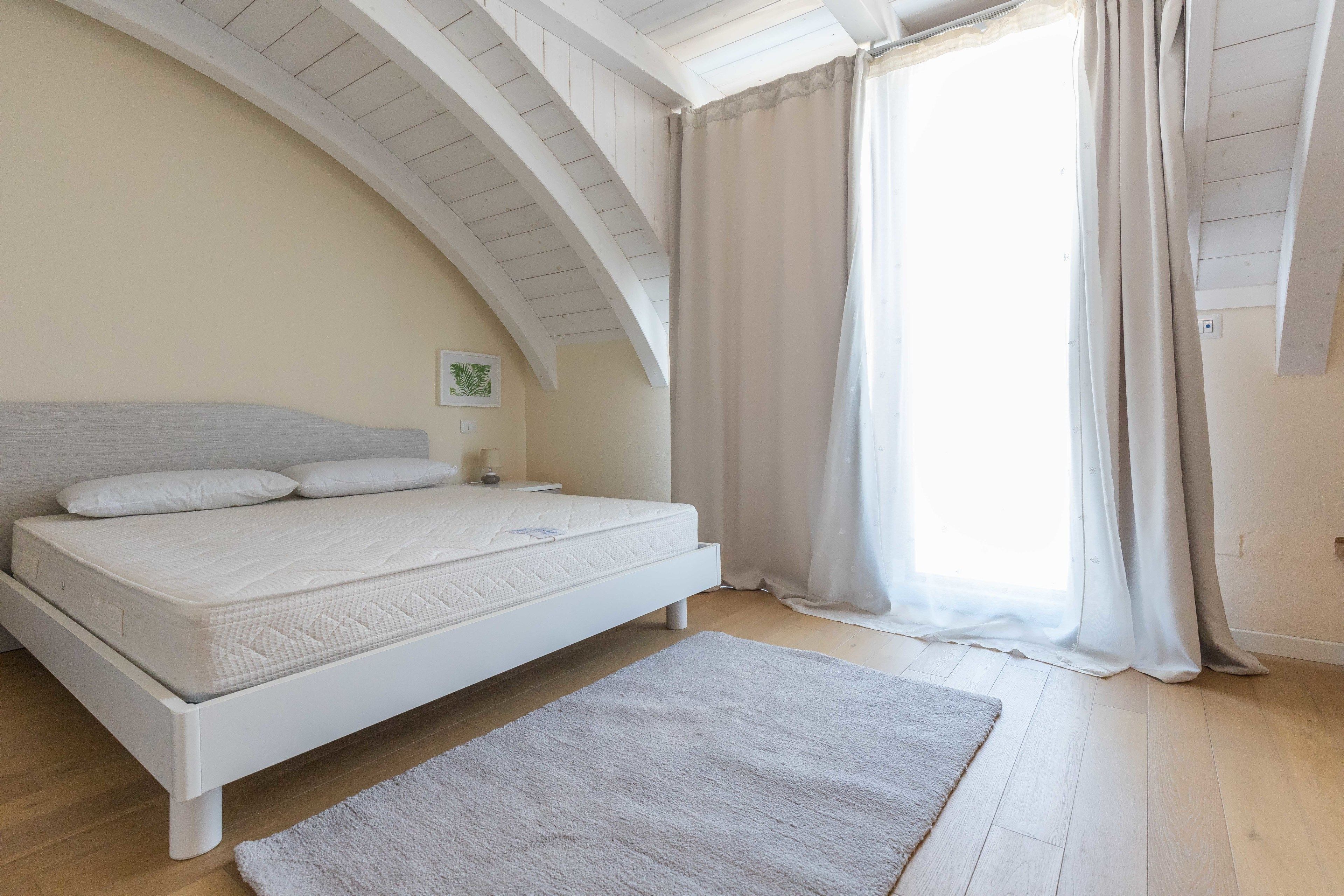 Un dormitorio con una cama blanca, cabecera blanca, mesita de noche blanca, pared blanca y una ventana con cortinas blancas, además de un suelo de madera.