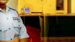 Ein Wachmann in Uniform mit Namensschild steht in einem Raum mit Wandverkleidung und einem Tisch mit einem Computermonitor.