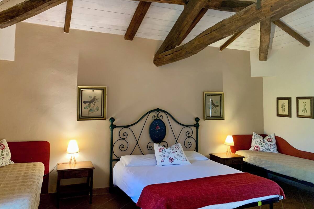 A bedroom with a wooden ceiling, a bed with a red blanket, two bedside tables with lamps, and framed pictures on the walls.