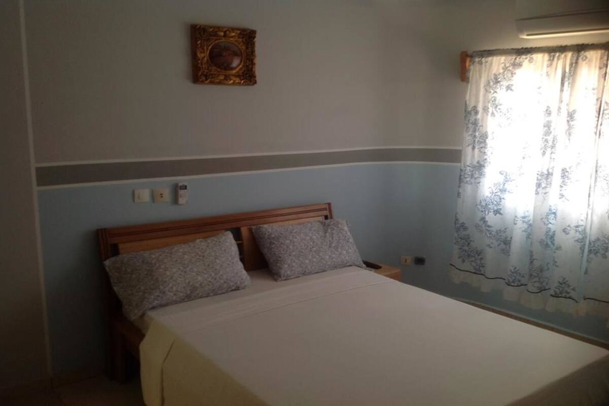 Een slaapkamer met een bed, een nachtkastje, een lichtschakelaar aan de muur en een raam met een bloemenprint gordijn.