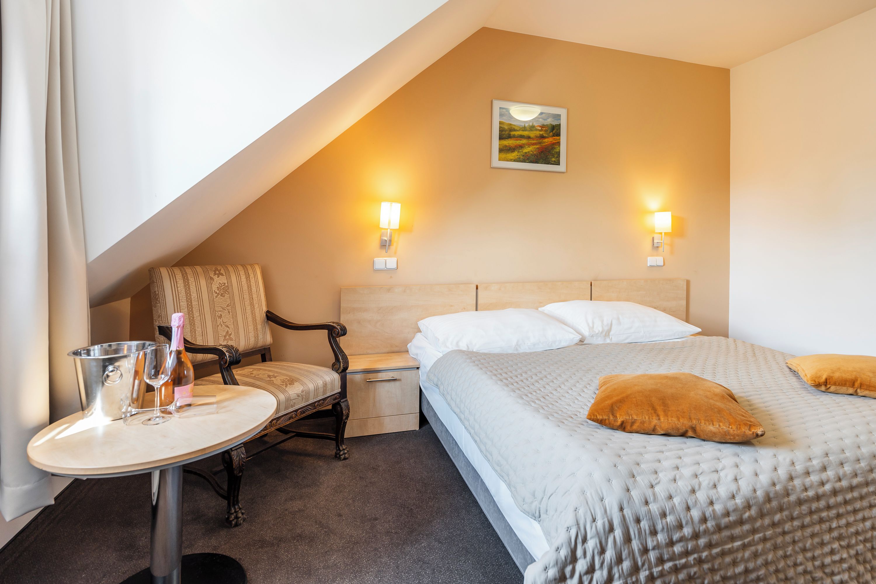 Ein Hotelzimmer mit einem Bett, Nachttischen, einem Sessel und einem kleinen Tisch mit Flasche und Glas.