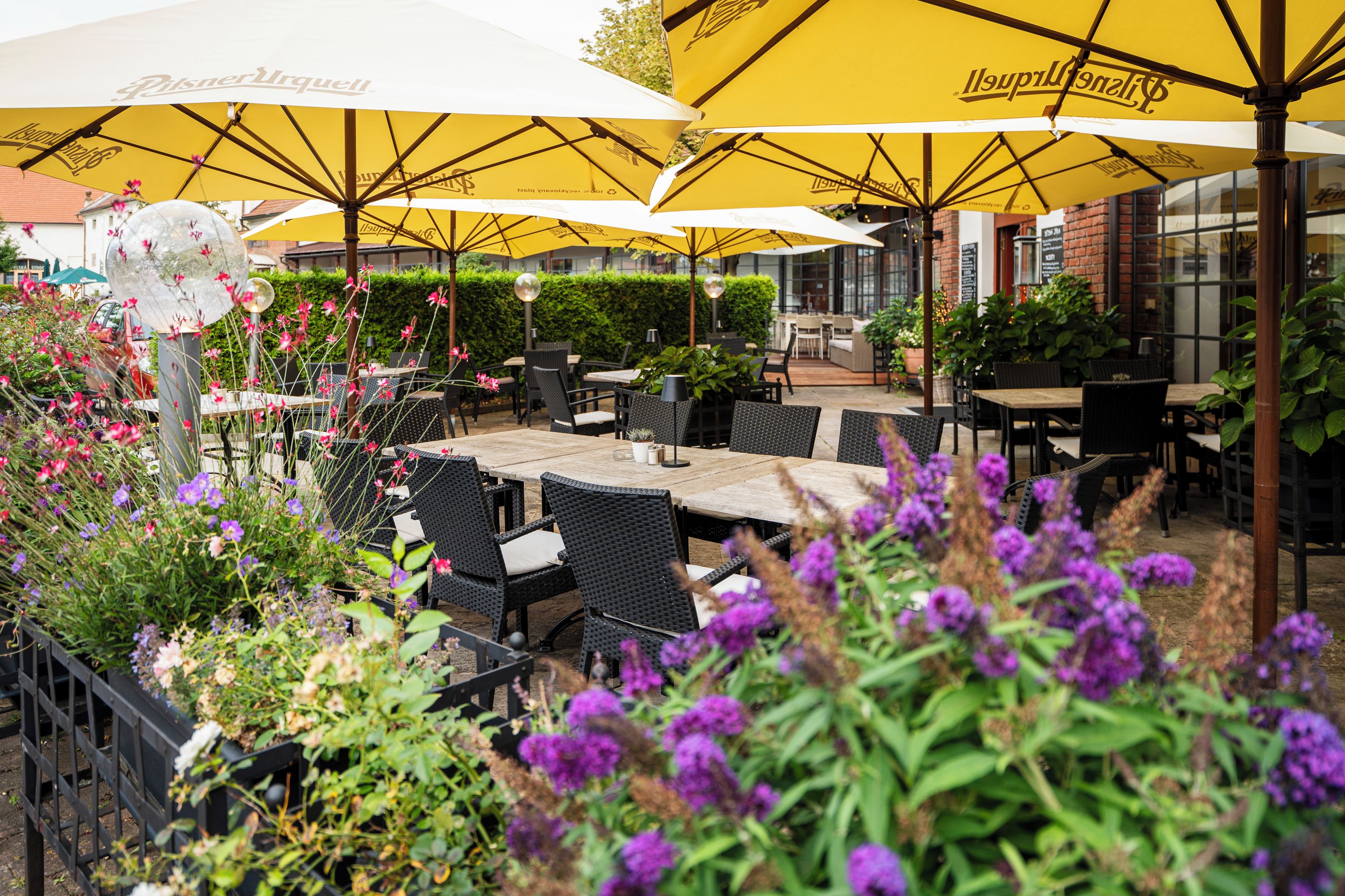 An outdoor seating area with tables and chairs under yellow umbrellas, surrounded by greenery and flowers.