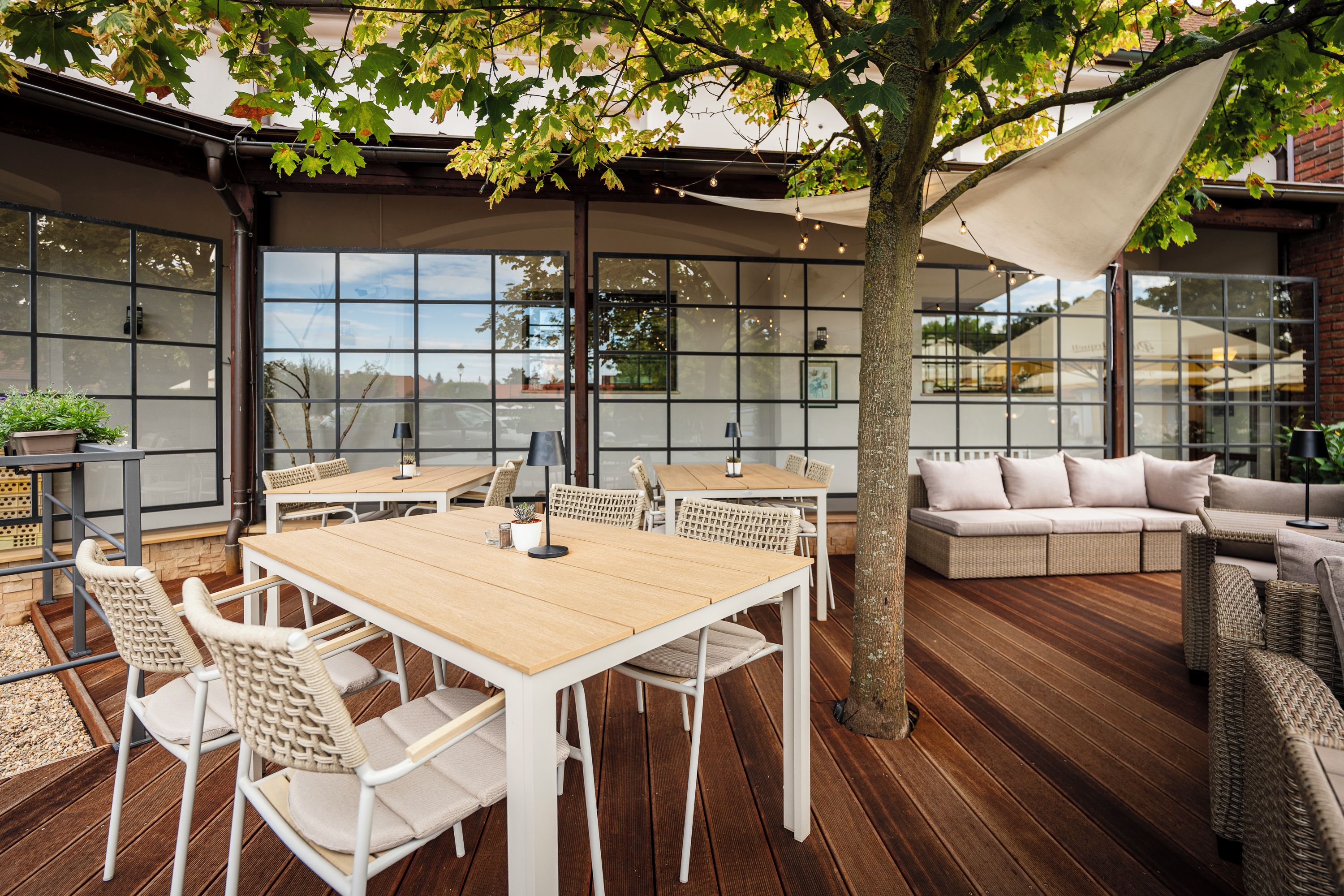 An outdoor patio with wooden tables and chairs, a cushioned seating area, and a large window overlooking a street.