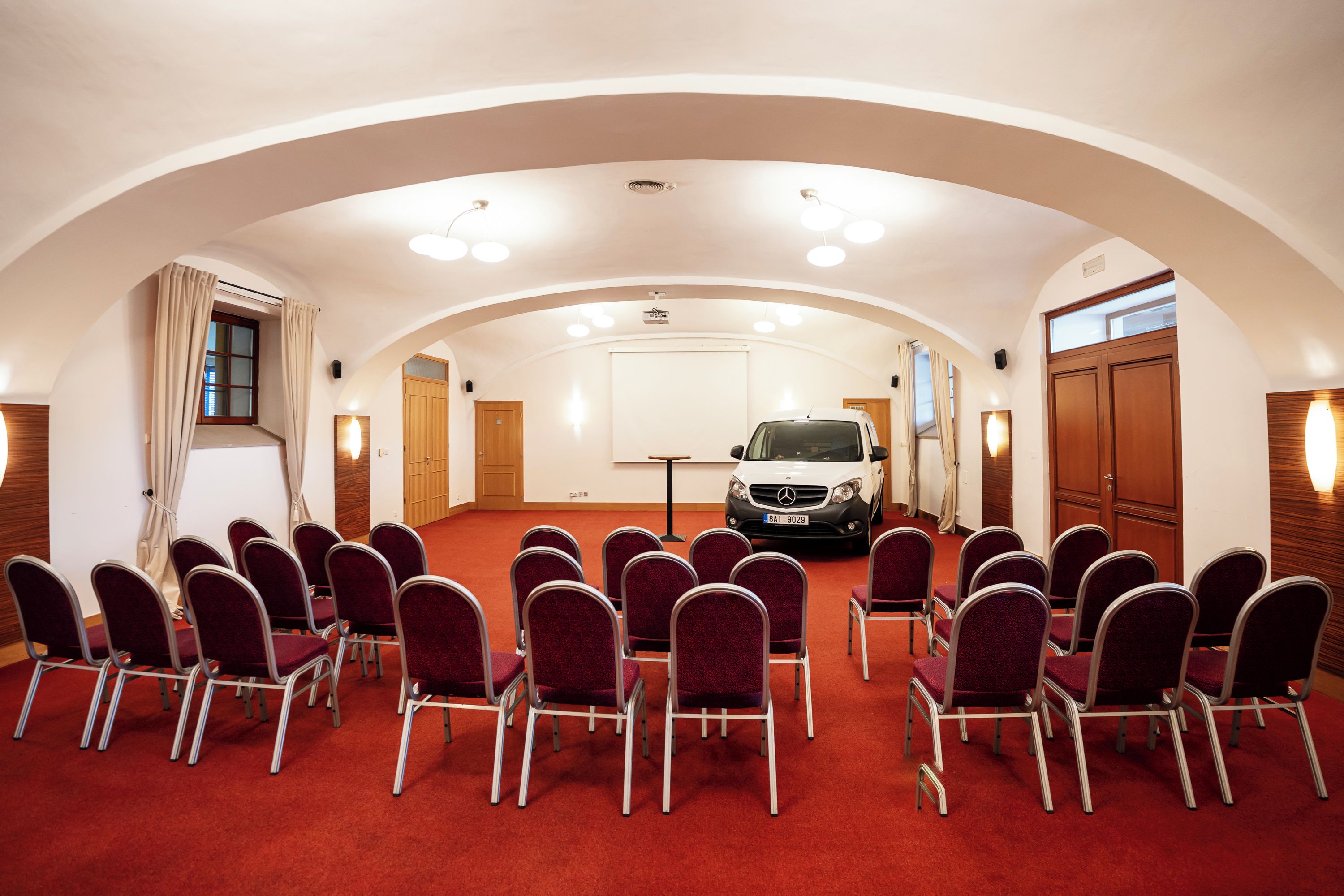 A conference room with a red carpet, rows of chairs, a white van, and wooden doors.