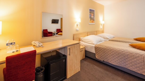 Ein Hotelzimmer mit Schreibtisch, Telefon, Mülleimer, einem Bett mit zwei Kissen und einem an der Wand montierten Fernseher.