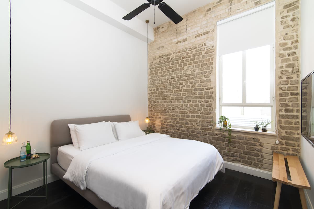 Quarto com parede de tijolos, cama com roupa de cama branca, mesa pequena com garrafa e copos, ventilador de teto e janela com plantas.