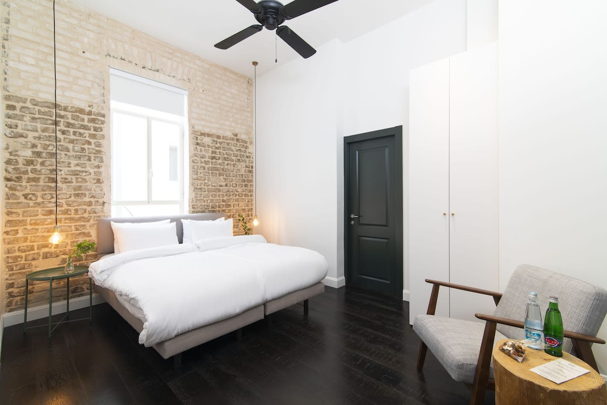 Quarto com parede de tijolos, uma cama, uma cadeira, uma mesa de madeira e uma porta.