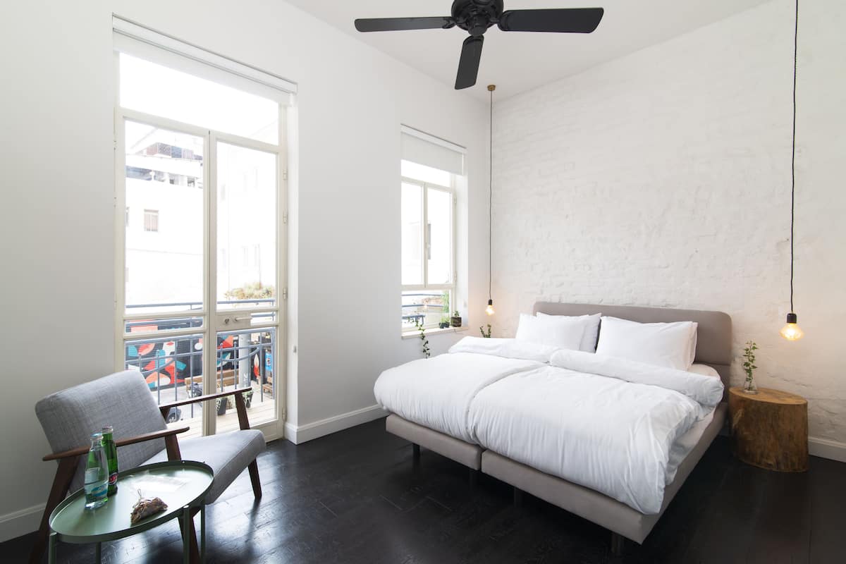 Quarto com cama grande, cadeira, mesa pequena, ventoinha no teto e janela com vista para o exterior.