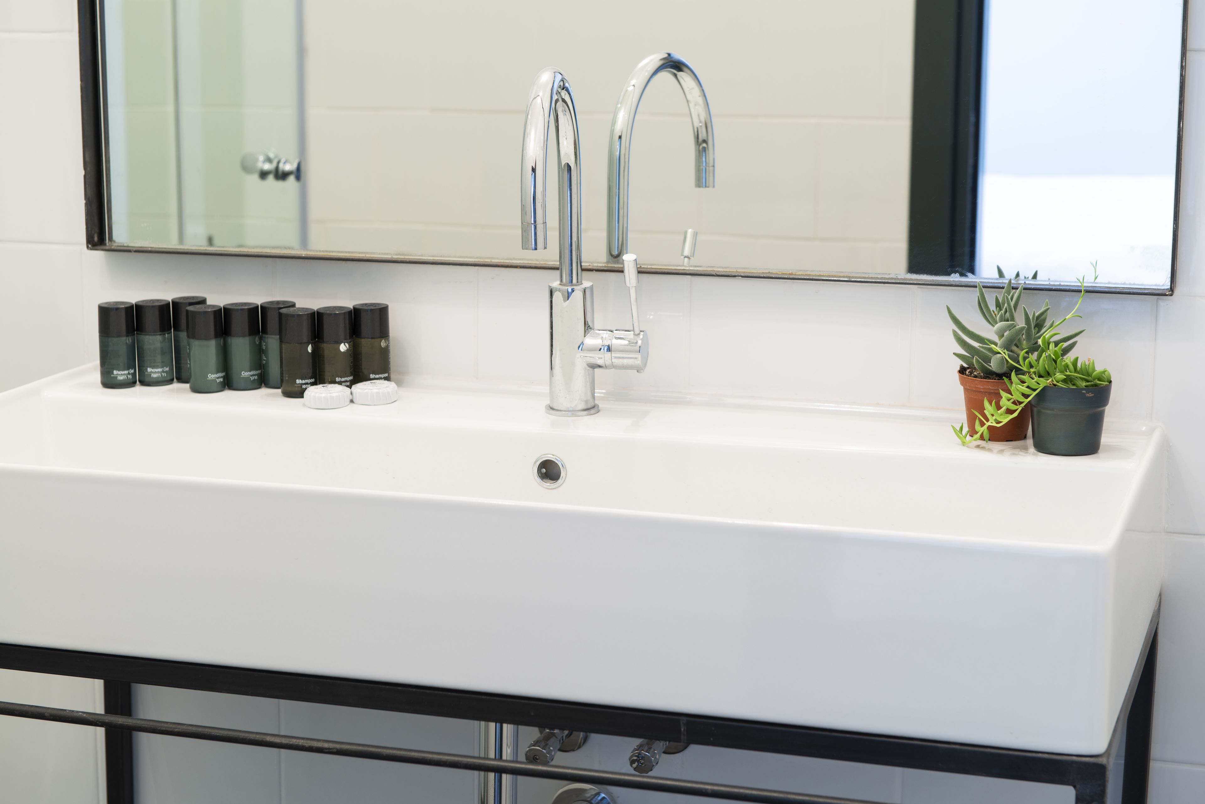 A white bathroom sink with two chrome faucets, a mirror above, and a row of toiletries on the left.