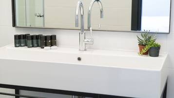 A white bathroom sink with two chrome faucets, a mirror above, and a row of toiletries on the left.