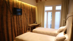 A hotel room with two beds, a flat-screen TV mounted on a brick-patterned wall, a small desk with a chair, and a window with curtains. - Muralha Charm House (Lamego)