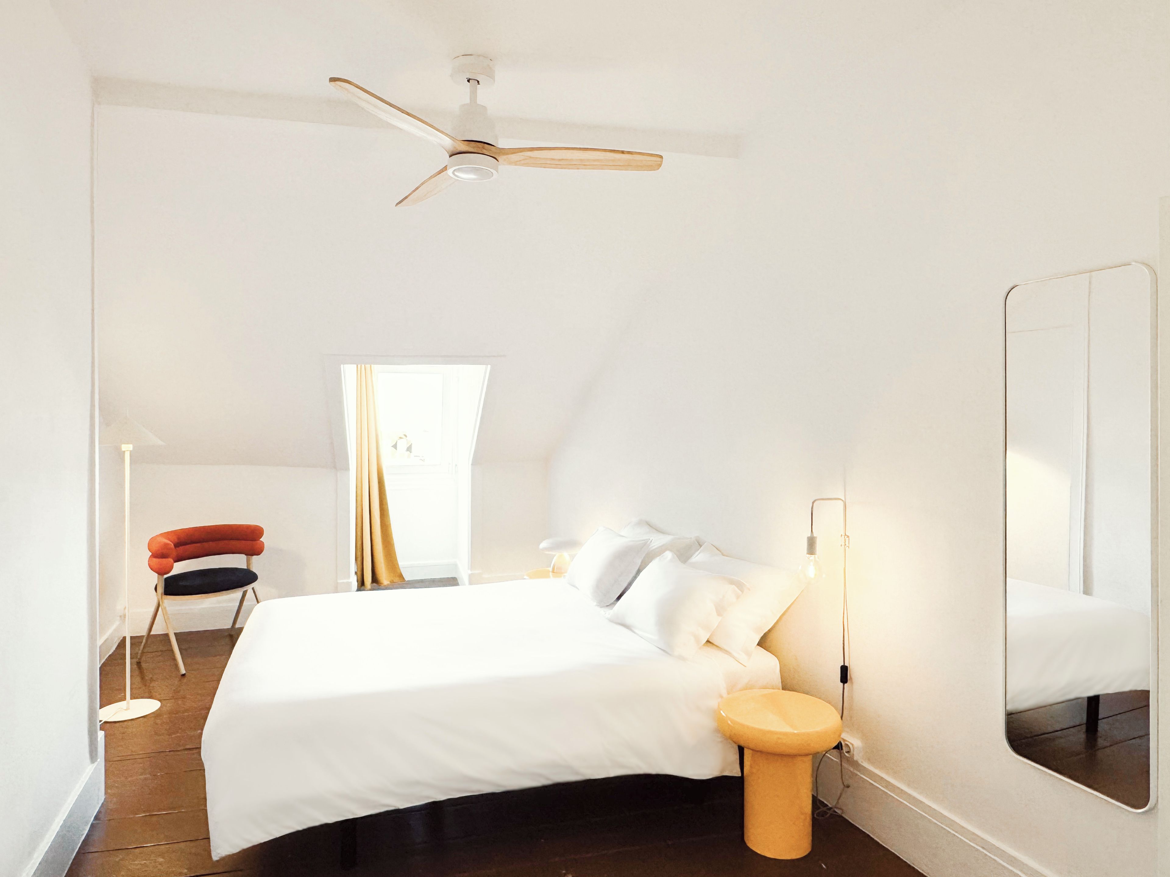 A bedroom with a bed, a bedside table, a mirror, a ceiling fan, and a window with curtains.