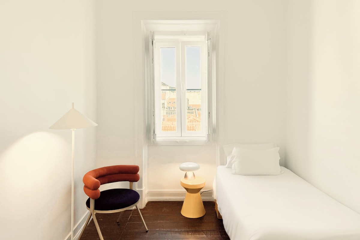 Une petite pièce lumineuse, avec un lit simple, une chaise, une lampe et une fenêtre donnant sur des immeubles.