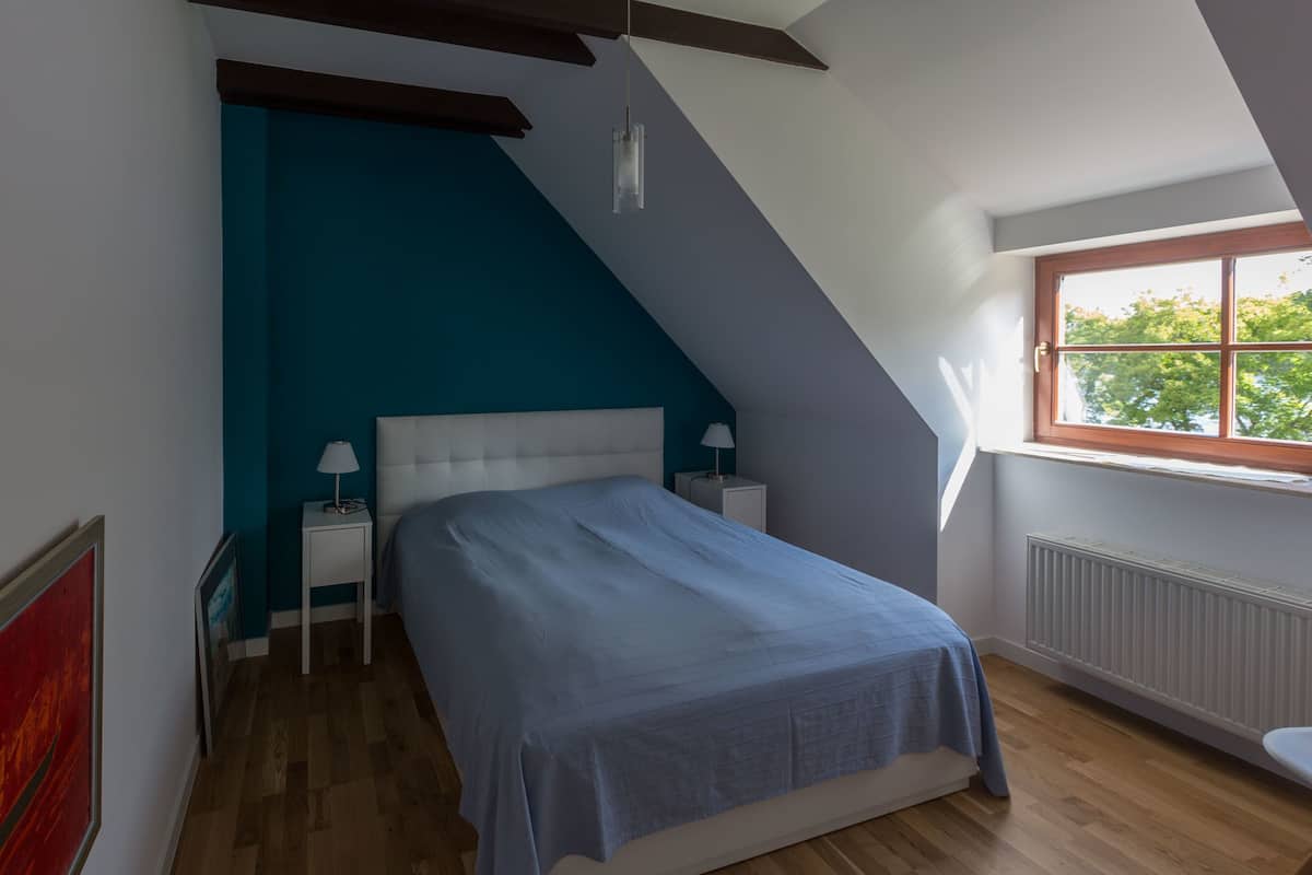 Ein Schlafzimmer mit schrägen Wänden, einem Bett mit blauer Bettdecke, zwei Nachttischen mit Lampen und einem Fenster mit Blick auf Bäume.