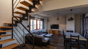 A modern living space with a wooden staircase, a sofa, a dining table with chairs, and a TV mounted on a brick wall. - ApartView - Osada Zamkowa Pasym (Pasym)