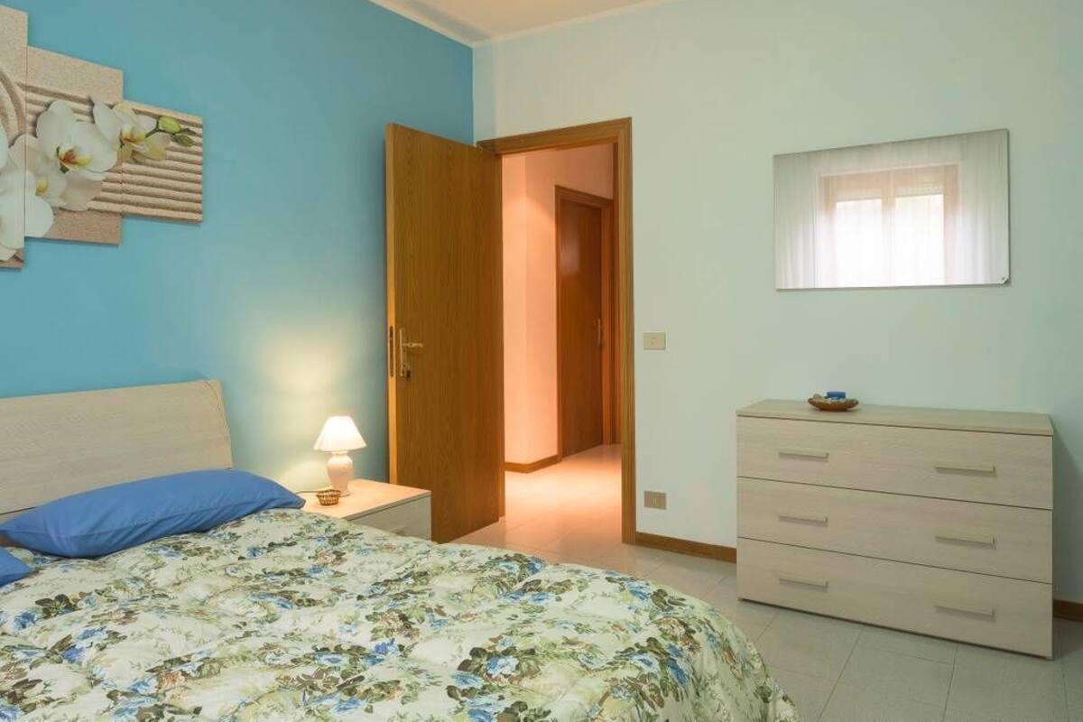 A bedroom with a bed, a nightstand, a dresser, a door leading to another room, and a window.
