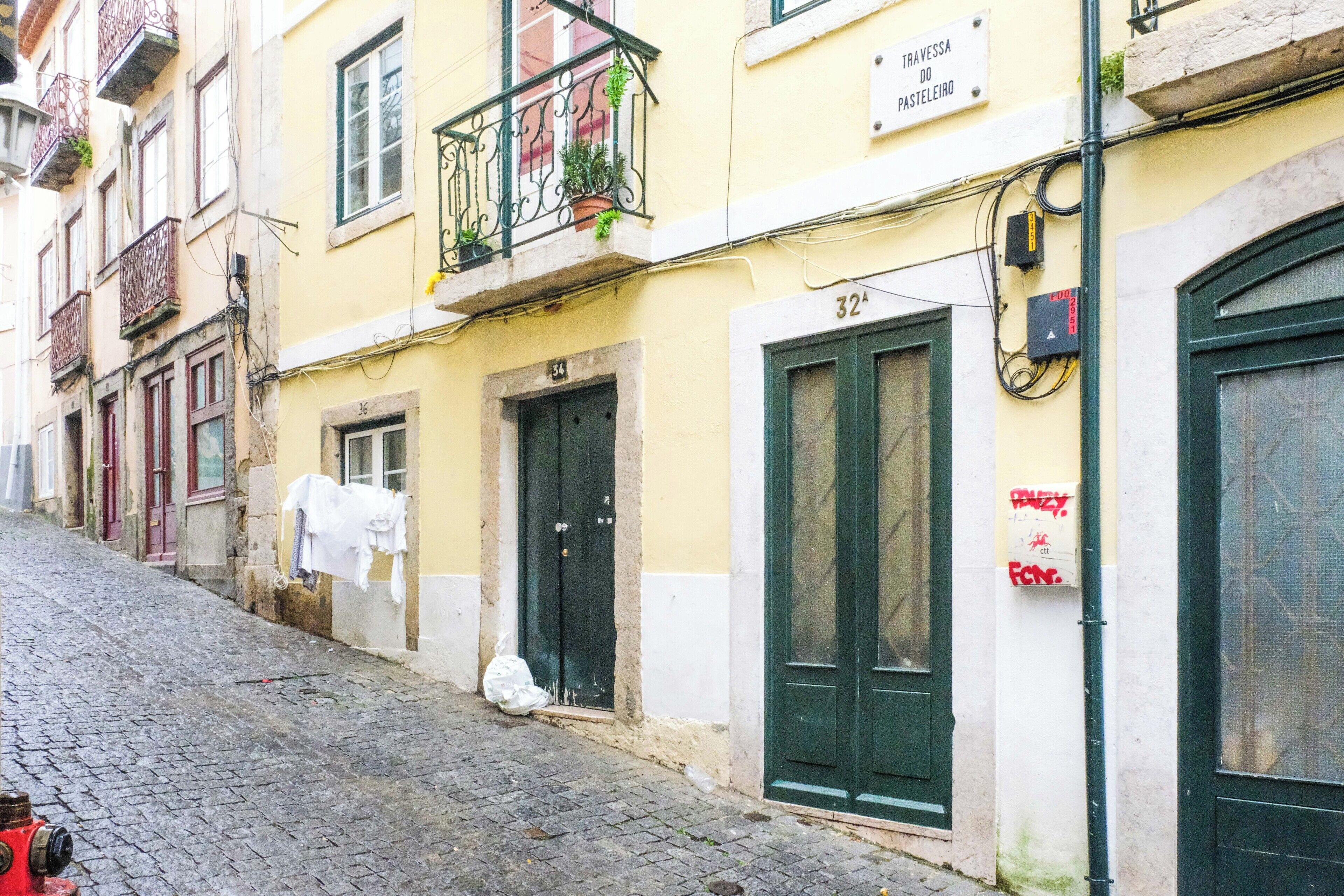 A narrow cobblestone street with buildings featuring balconies with potted plants, a visible fire hydrant, and a sign indicating a pasteleria.