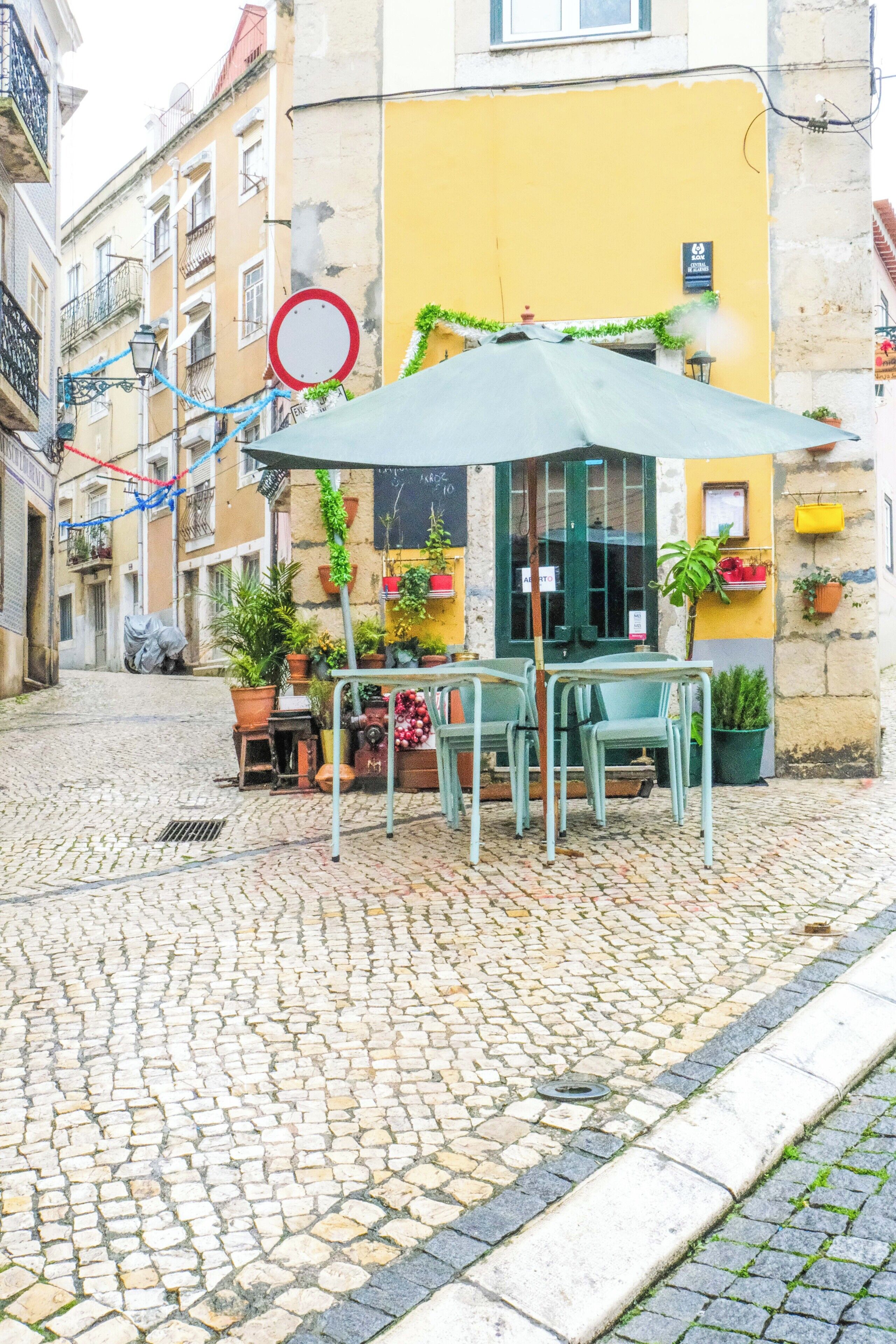 En kullerstensgata med ett cafébord och stolar under ett grönt parasoll, omgivet av planterade blommor och en gul byggnad.