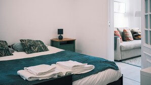 A neatly made bed with a teal blanket, a bedside table with a lamp, and a view into a living area with a sofa and colorful pillows. - 03 Nice Flat by Travessa do Pasteleiro (Lisbon)