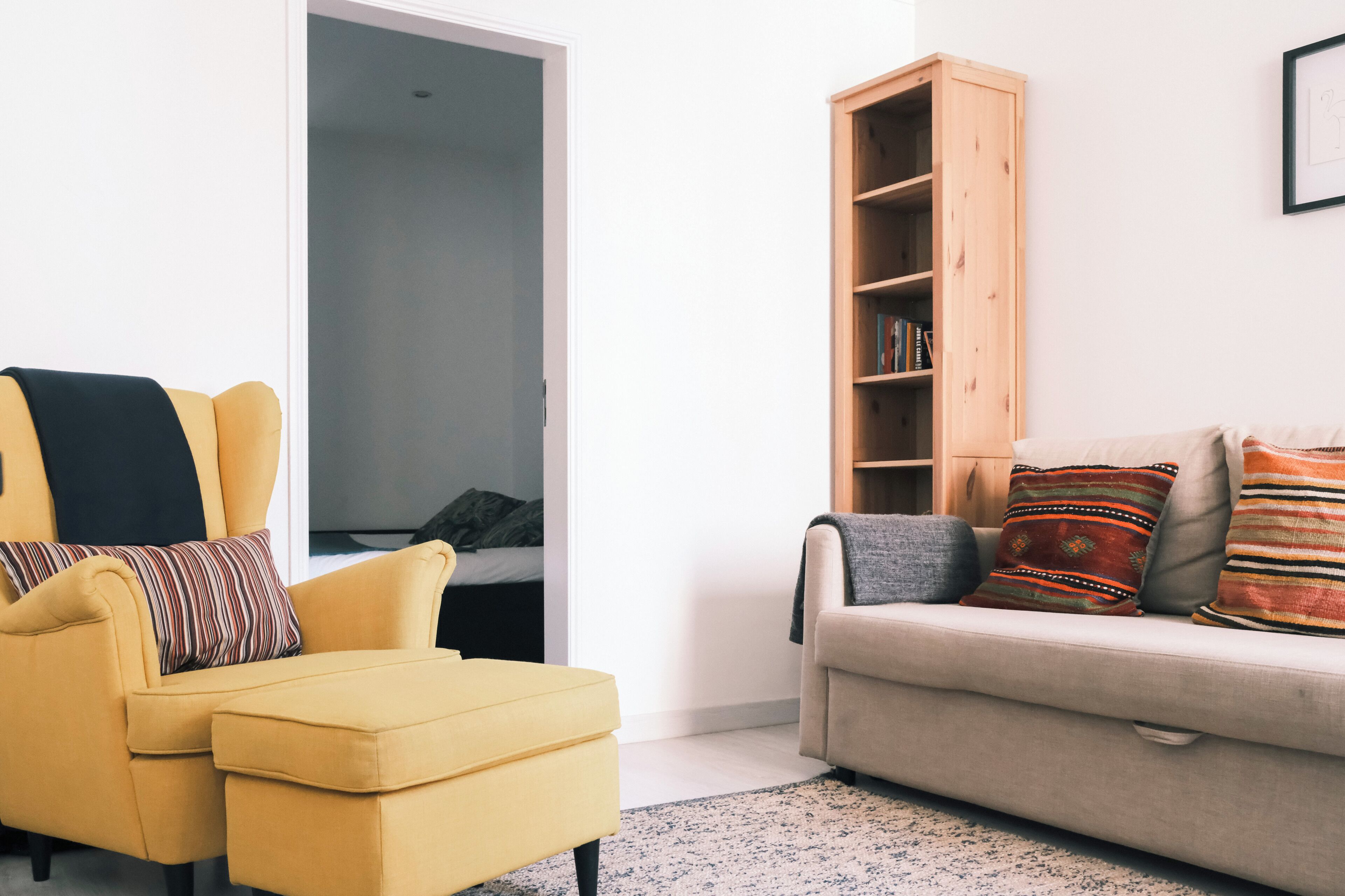 A living room with a yellow armchair, a beige sofa, a wooden bookshelf, and a framed picture on the wall.