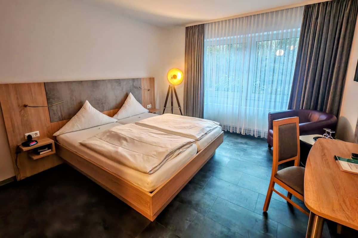 A hotel room with a wooden bed, a bedside table with a lamp, a chair, and a table.