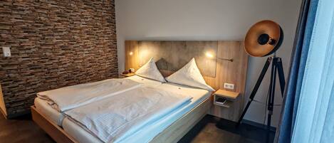 Un dormitorio moderno con pared de piedra, cabecera de madera, cama con ropa blanca, mesita de noche con lámpara y lámpara sobre trípode.