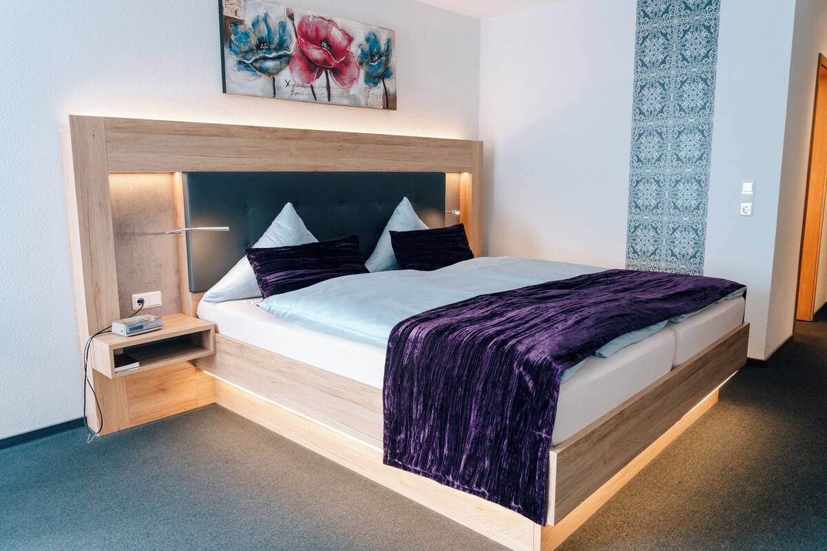 A modern hotel room with a wooden headboard, a flat-screen TV, a bedside table with a power outlet, and a large window.