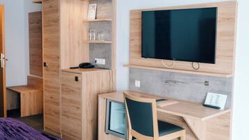 A compact room with a wooden desk, a chair, a television mounted on the wall, and a wardrobe with shelves.