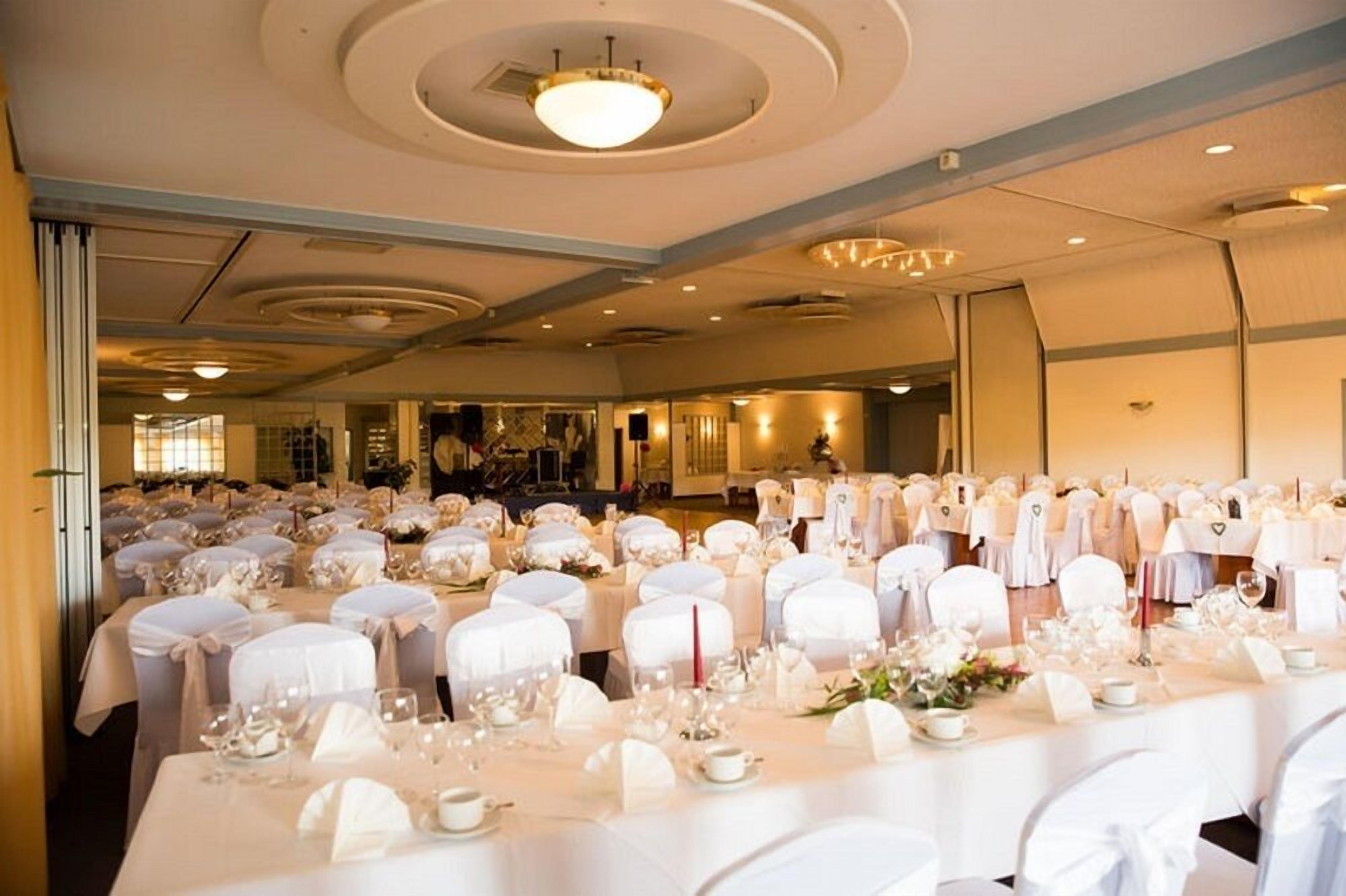 Uma sala de banquetes com mesas redondas preparadas para um evento formal, com toalhas brancas e capas de cadeiras combinando.