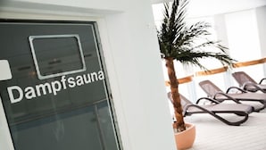 Señal de Dampfsauana en una puerta de vidrio, una palmera en maceta y sillones reclinables en un espacio interior luminoso.