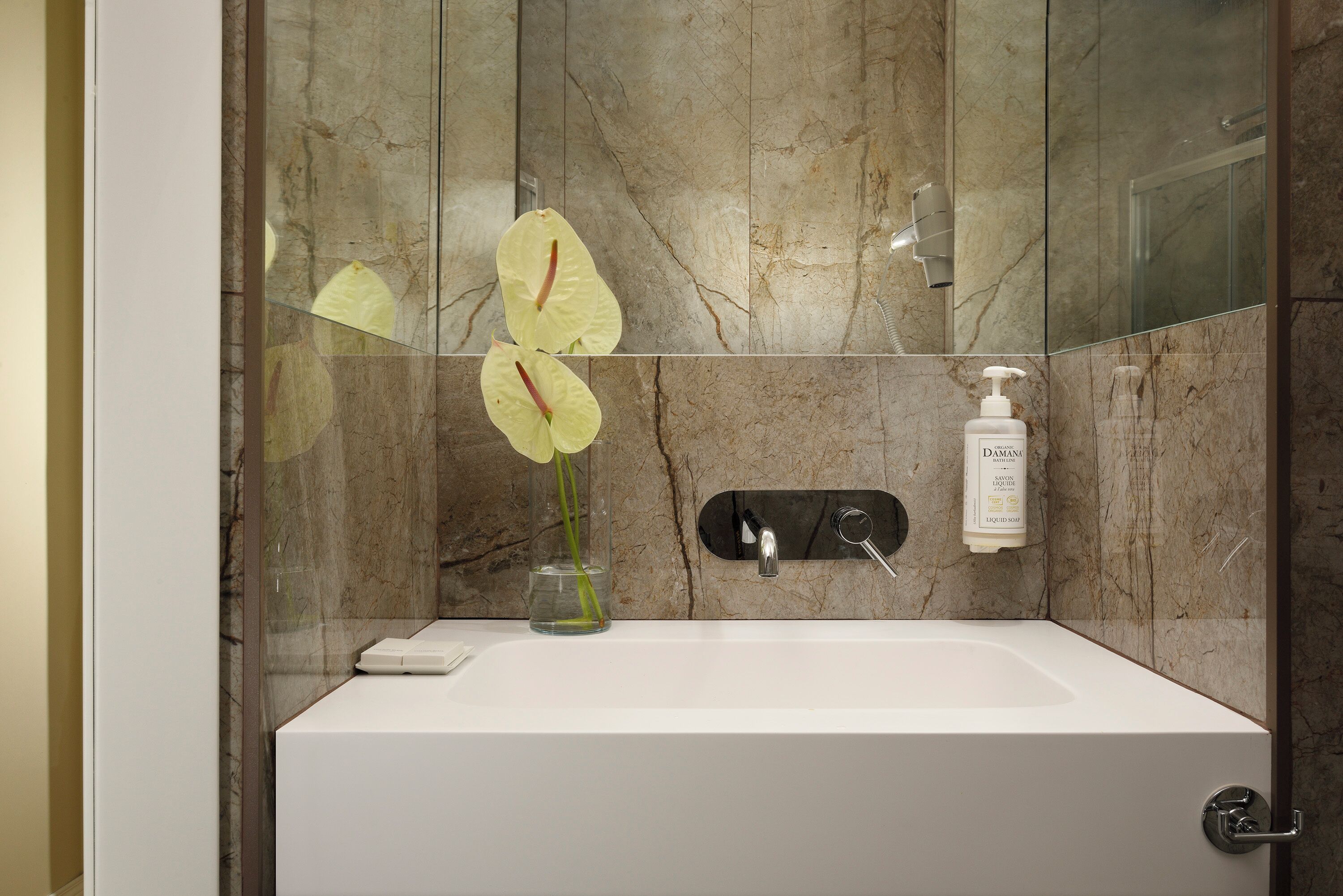 A modern bathroom with a marble wall, a white sink, a glass vase with flowers, and a bottle of shampoo.