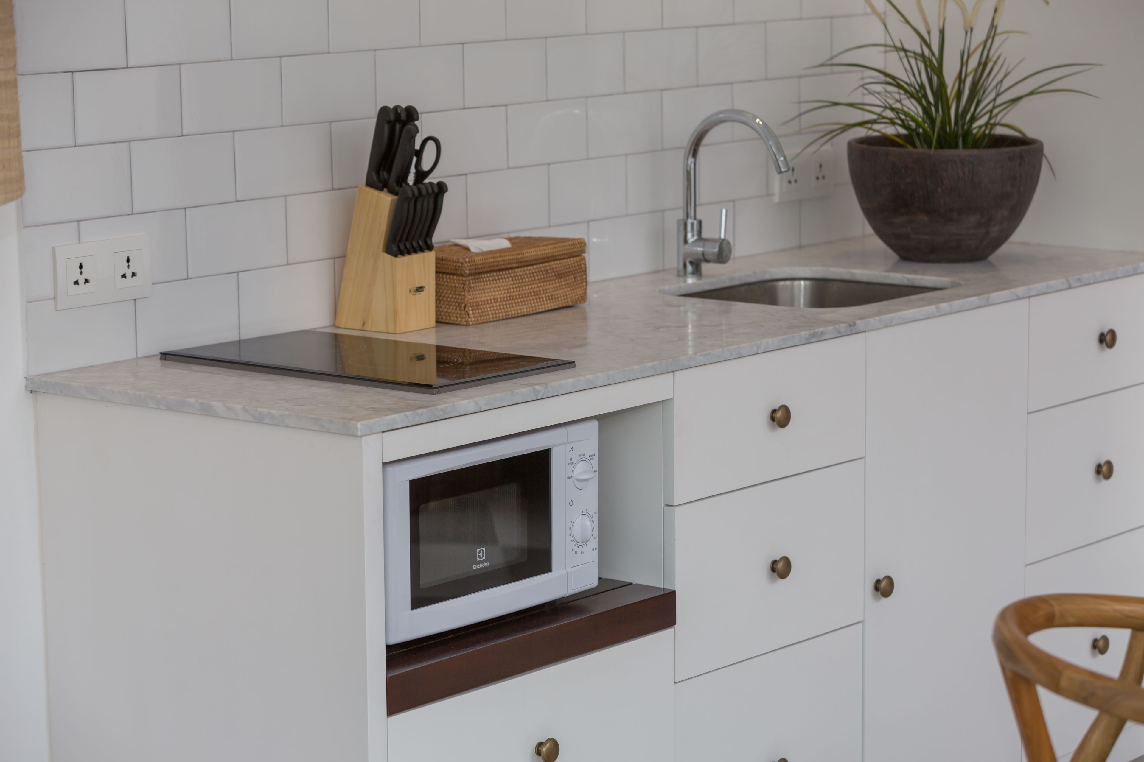 Una cocina con armarios blancos, microondas integrado, un lavamanos, tabla de cortar de madera, bloque de cuchillos y una planta en maceta.