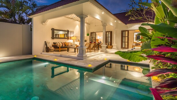 A well-lit outdoor pool area with a covered seating area, a dining space, and a view of the house. - Villa CJ (Seminyak)