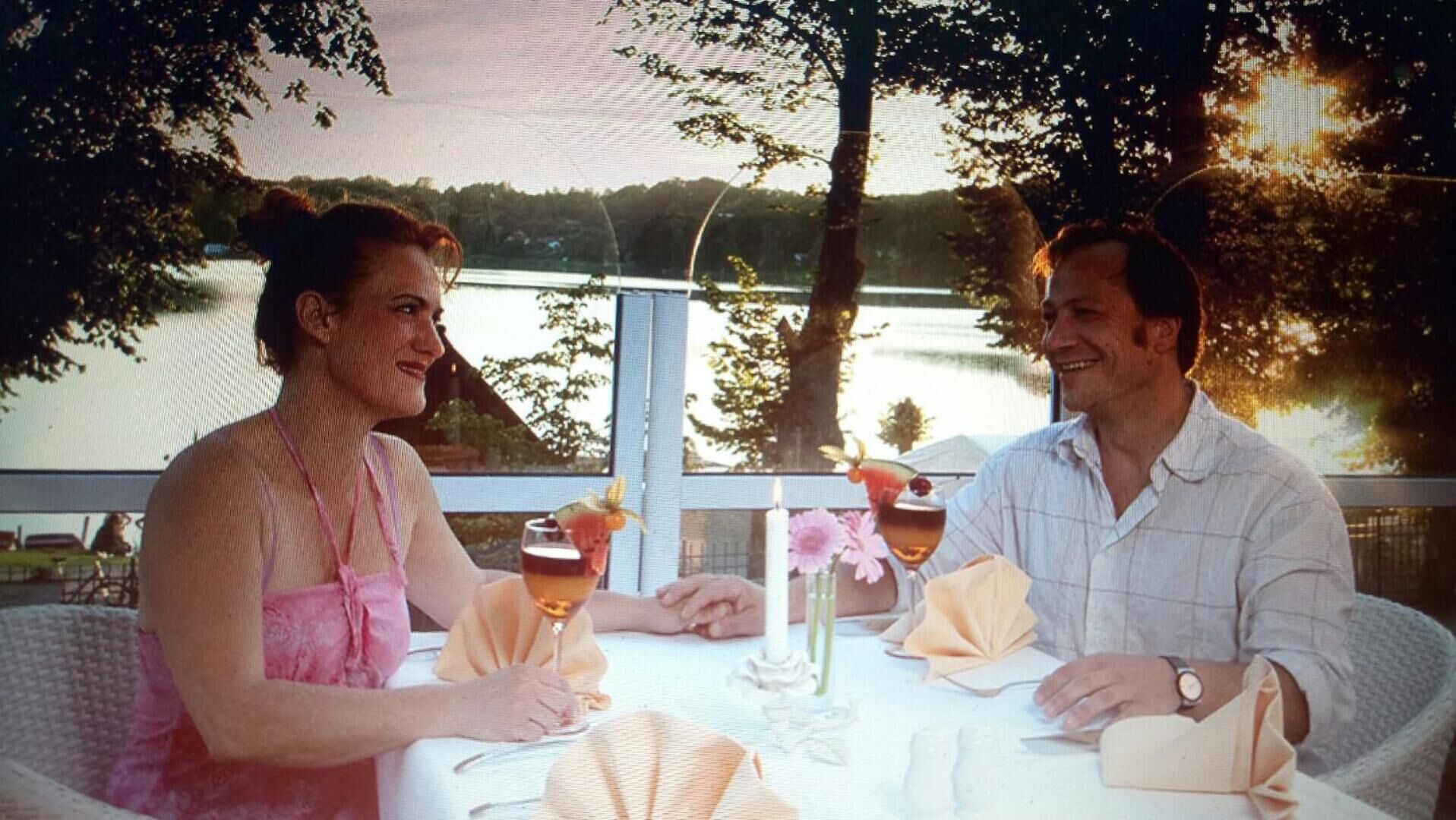 A couple dining at an outdoor table with a view of a lake, surrounded by trees.