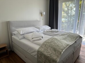 A neatly made bed with white linens and a patterned throw blanket, a bedside table with a lamp, and a view of trees through a window with curtains. - Strandhotel Buckow Resort & SPA (Buckow)