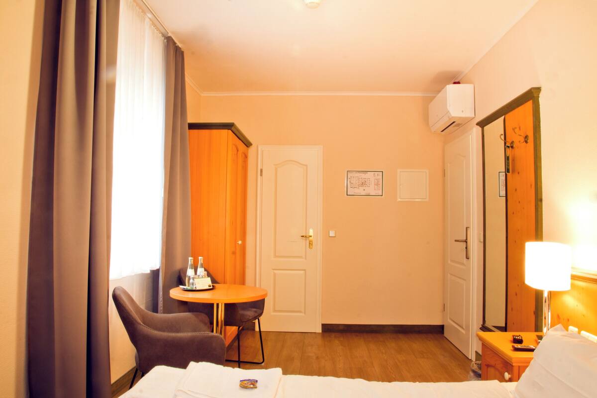 A hotel room with a bed, a chair, a small table with a bottle and glasses, a lamp, and a door.
