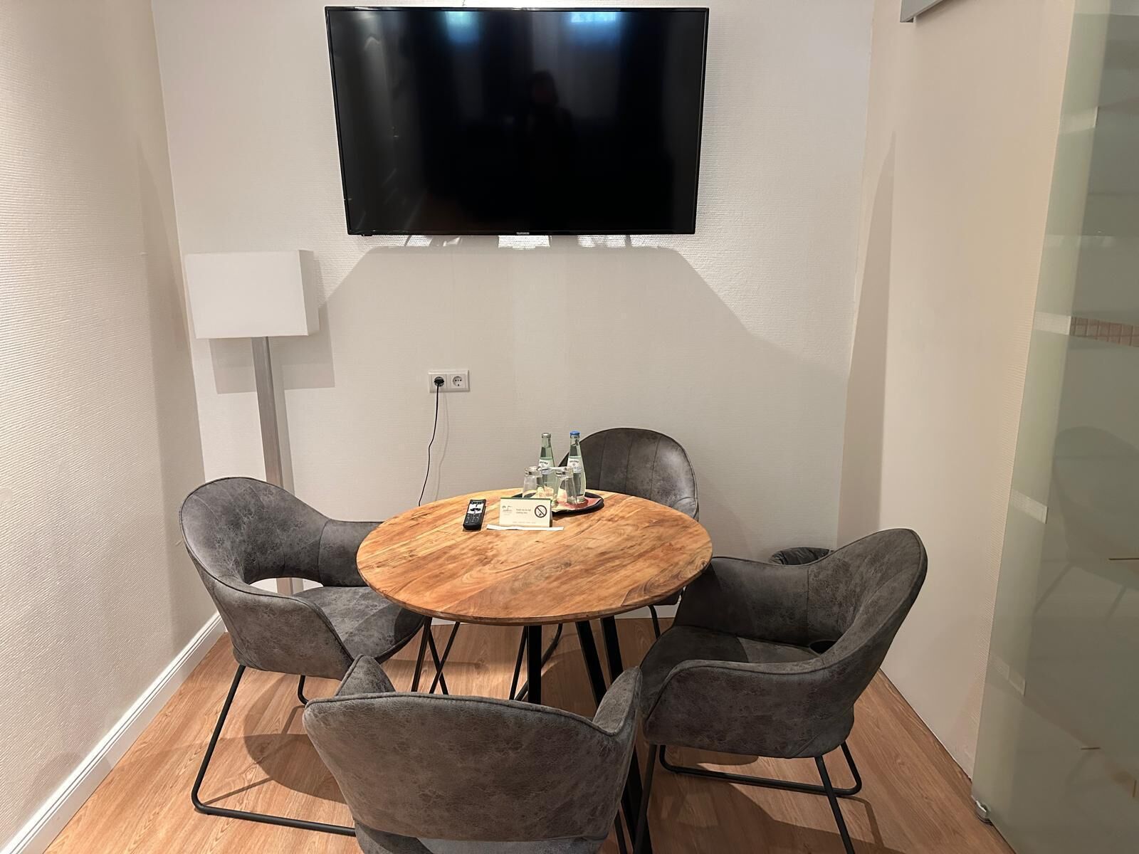 Un salón de conferencias con una mesa redonda de madera, cuatro sillas grises tapizadas, un televisor de pantalla plana montado en la pared y una pequeña mesa con un control remoto y un vaso de agua.