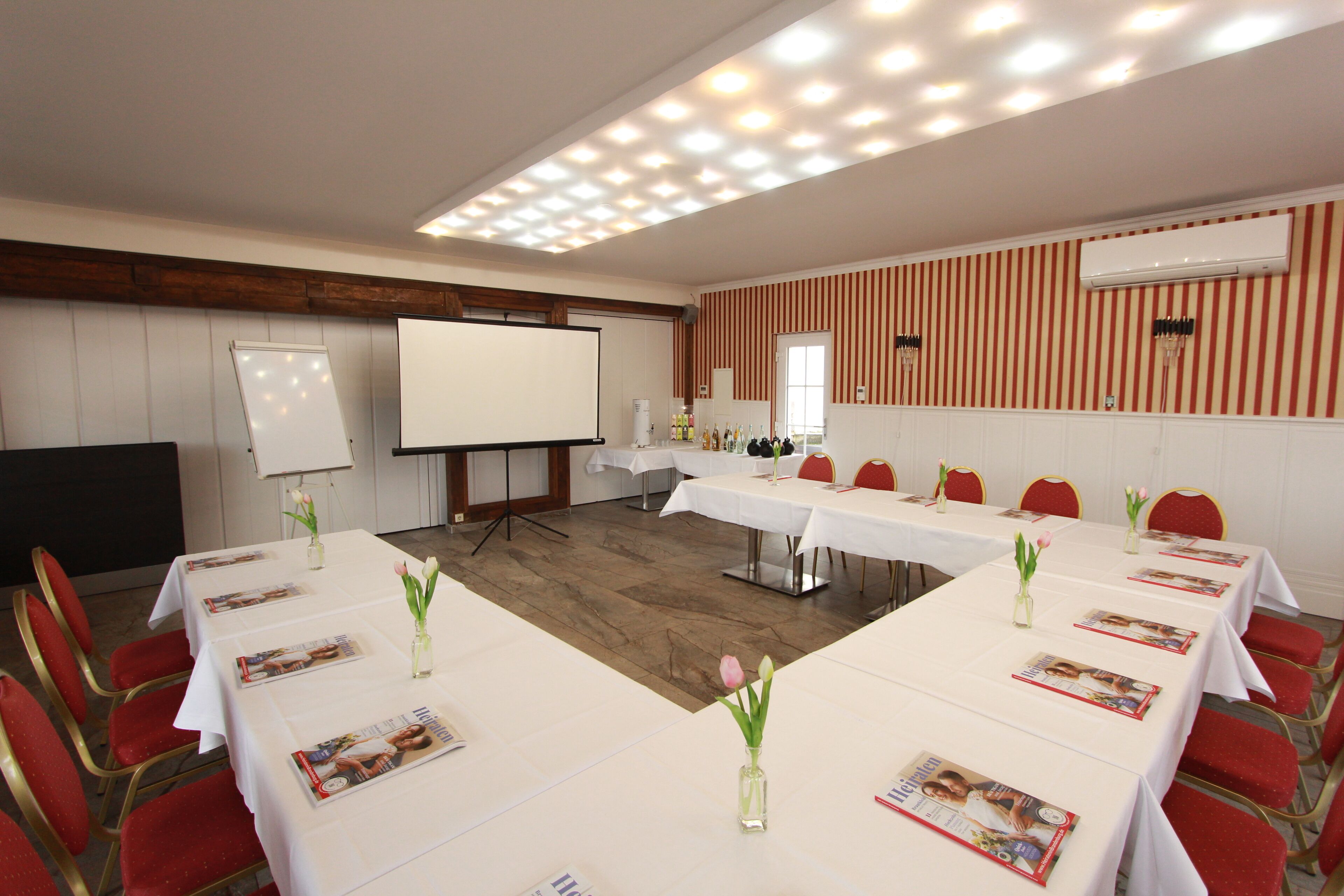 Un salón de conferencias con mesas blancas, sillas rojas y una pizarra.